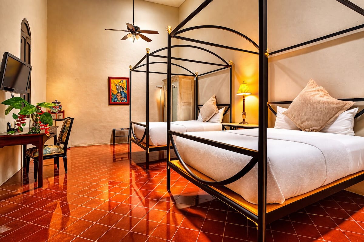 A room with a four-poster bed, a desk with a chair, a television, and a plant.