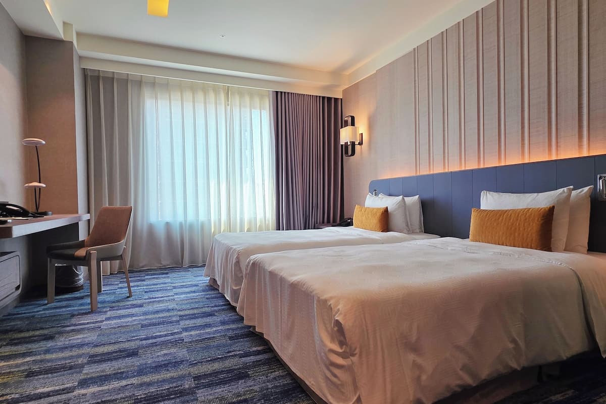 Een hotelkamer met twee bedden, een bureau, een stoel en een groot raam met gordijnen.