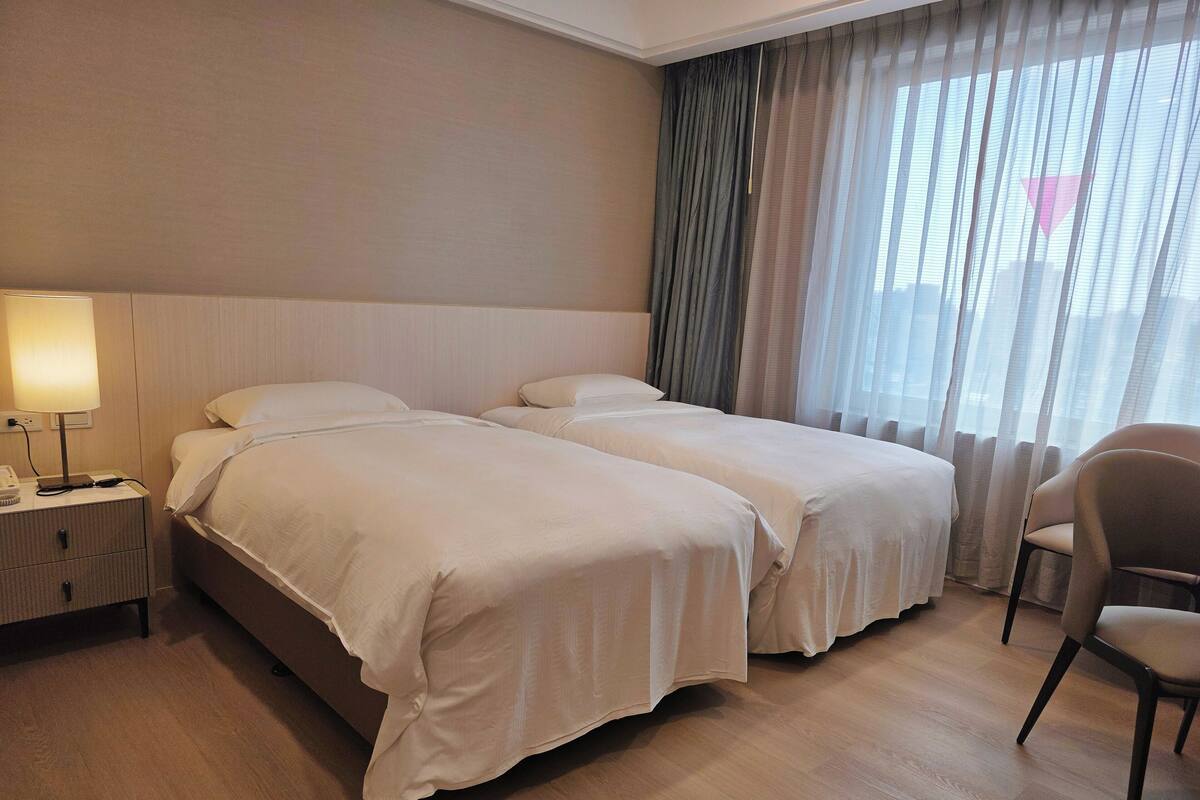 Een hotelkamer met twee bedden, een nachtkastje met een lamp, een stoel en een raam met gordijnen.