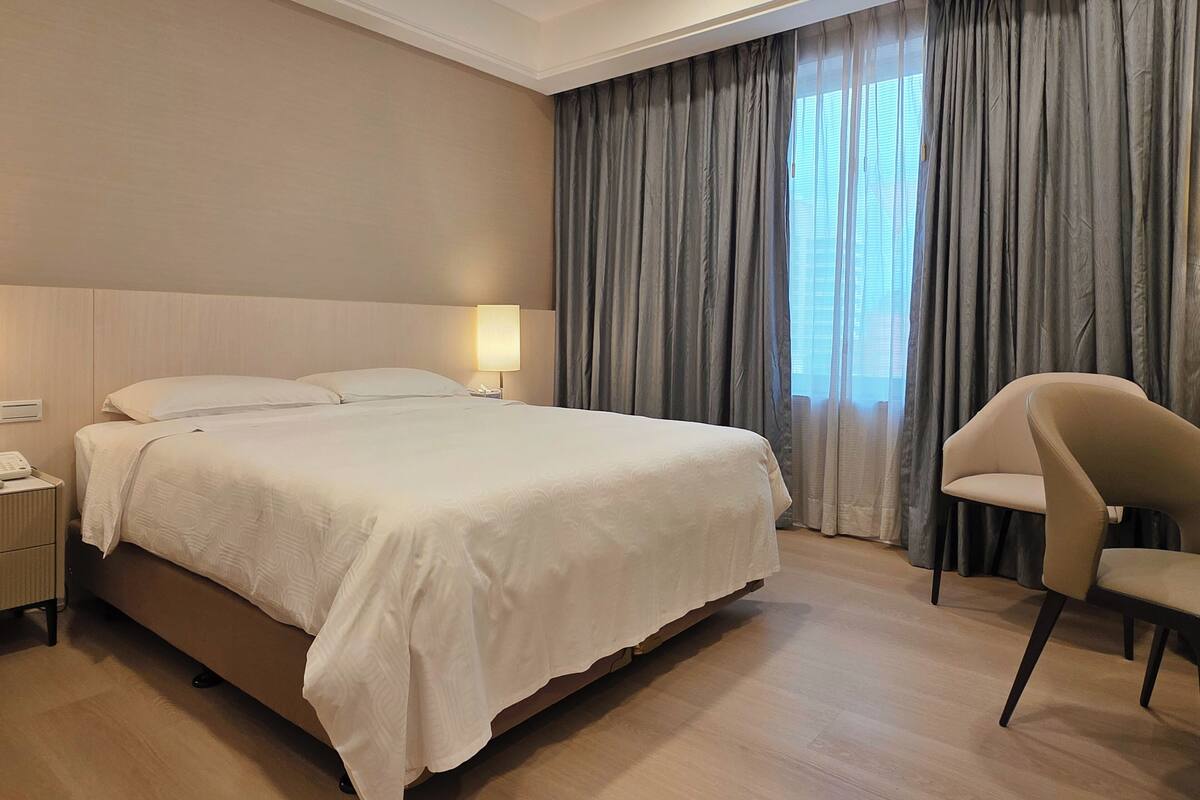 Een hotelkamer met een groot bed, een bedlamp, een stoel en een raam met gordijnen.