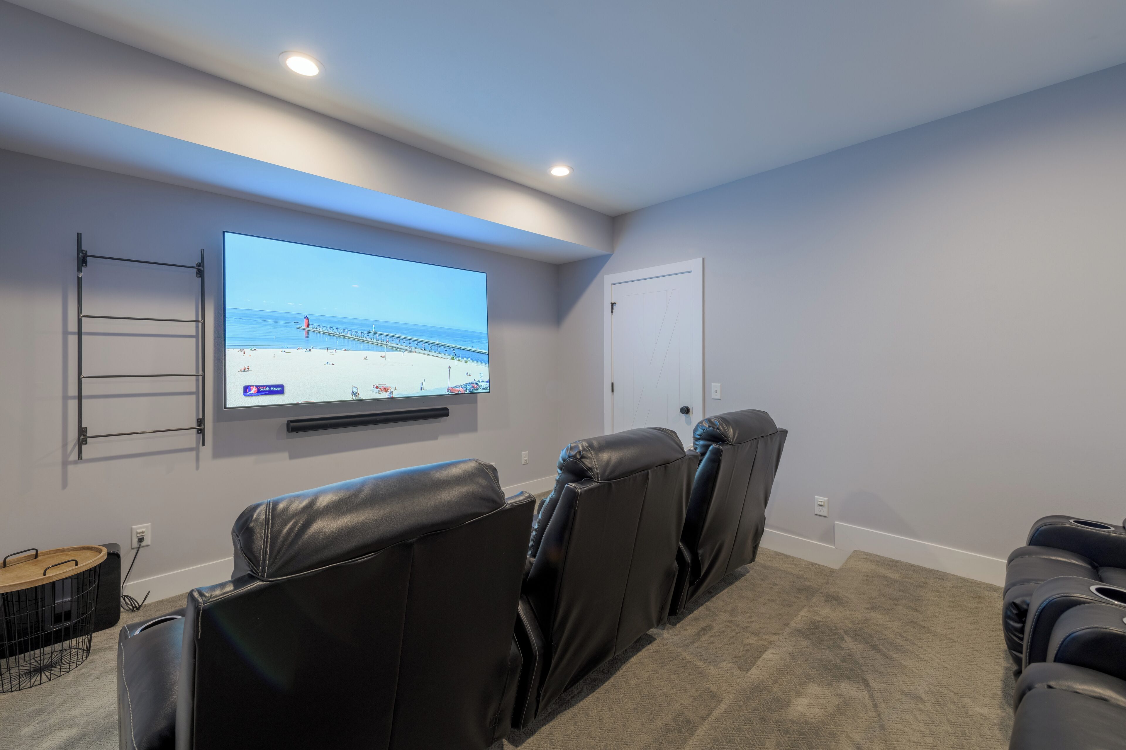 Un salón de cine en casa con un televisor de pantalla plana grande montado en la pared, sillones de cuero reclinables, una mesa de centro de madera y una puerta.