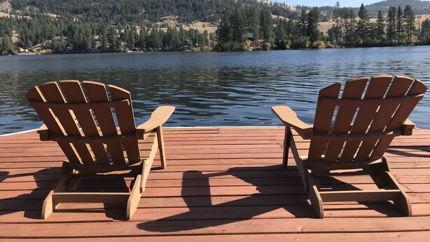 Due poltrone in legno di stile Adirondack su un pontile di legno con vista su un lago e un pendio boscoso sullo sfondo.