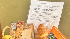 Una cesta de mimbre llena de pan, frutas y aperitivos empaquetados, con un aviso sobre la política de no recepción y las instrucciones para el registro sin contacto.