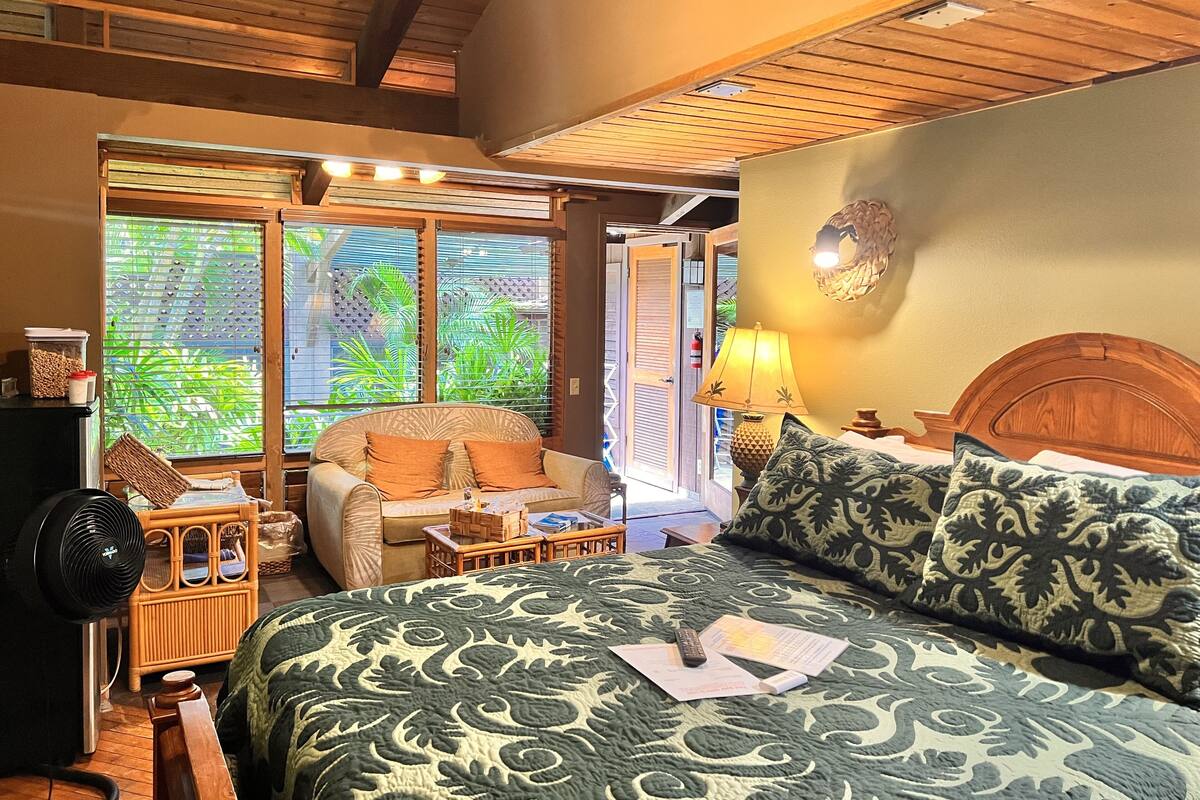 A cozy room with a bed, a chair, a table, and a fan.