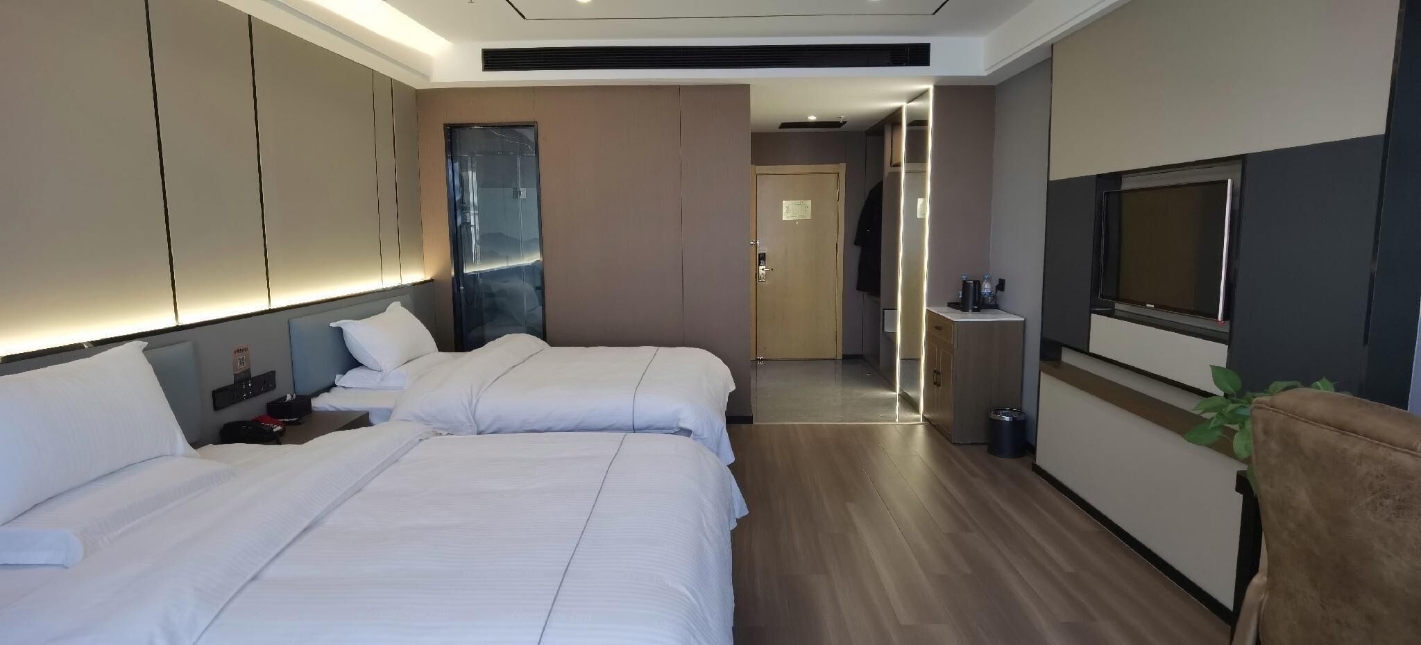 A hotel room with two beds, a TV, a small kitchenette, and a bathroom visible through an open door.