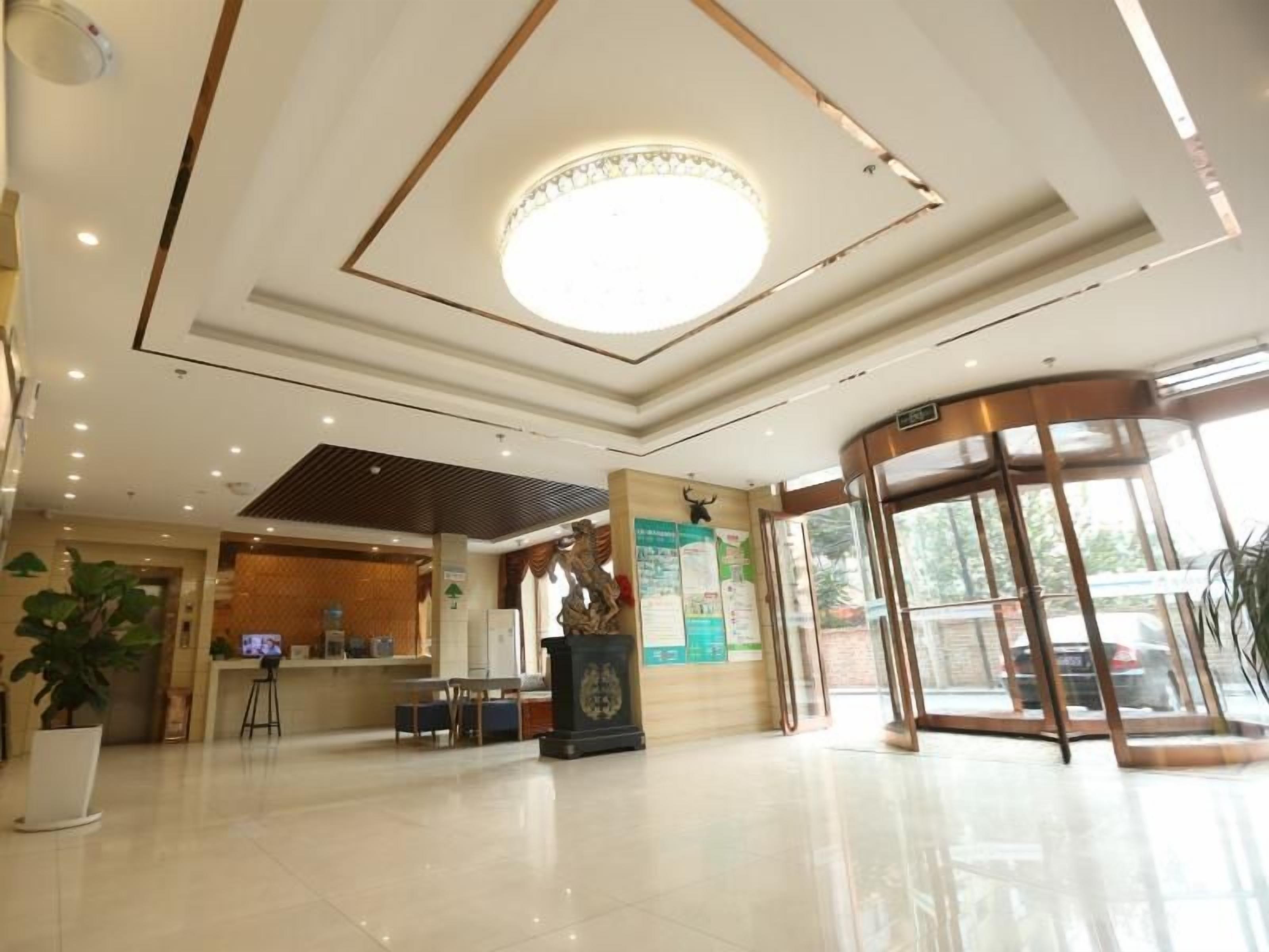A hotel lobby with a reception desk, a large circular light fixture, a glass elevator, and a potted plant.