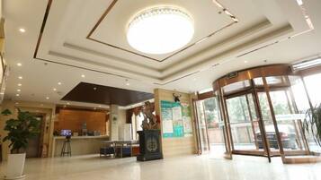 A hotel lobby with a reception desk, a large circular light fixture, a glass elevator, and a potted plant.