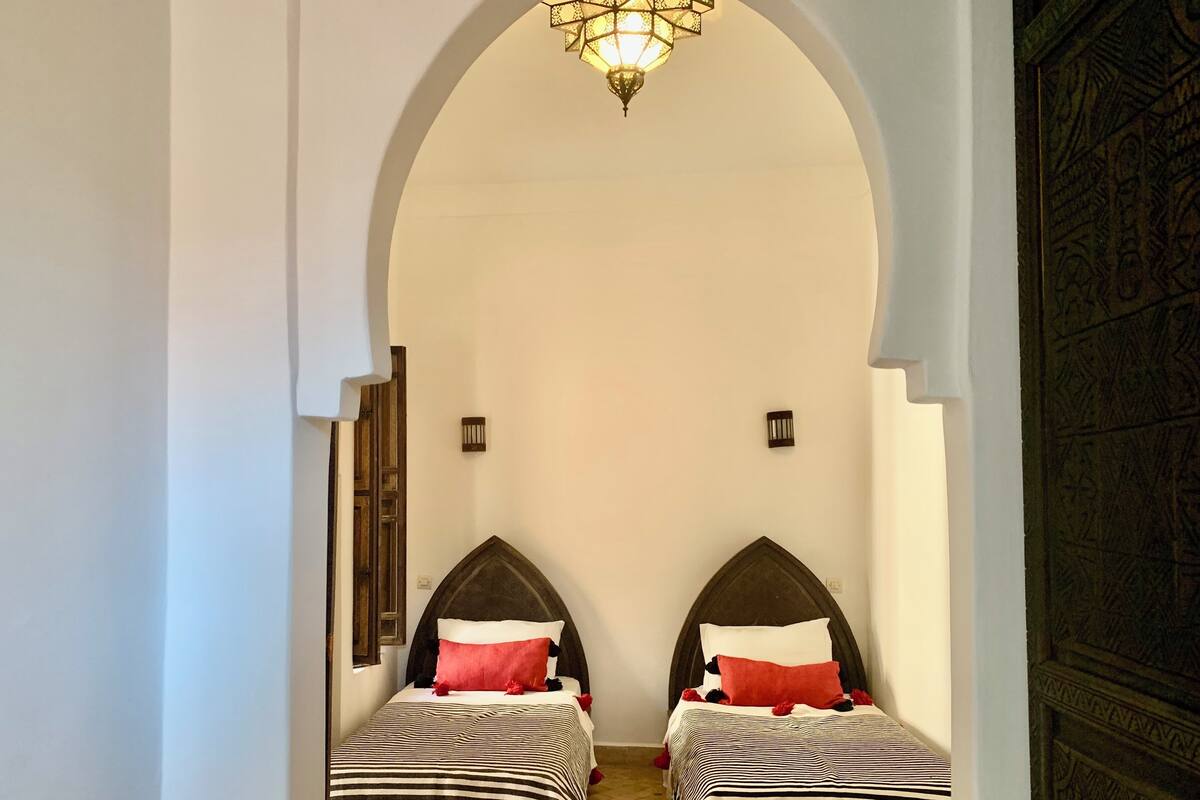 Une chambre avec deux lits, un tapis à motifs, un lustre et une porte en arc de cercle.