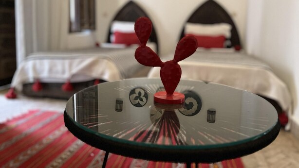 A glass table with a red object on top, a bed with white bedding and red pillows, and a wall with various circular decorations. - Riad Laksiba (Marrakech)