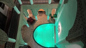 A pool with a green hue, surrounded by ornate columns and arches, and a seating area with a red cushion. - Riad Laksiba (Marrakech)