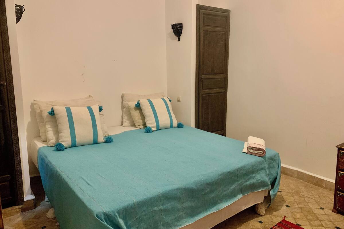 Un lit recouvert d’une couvre-lit bleue, deux oreillers à rayures bleues, une petite table de chevet avec une serviette blanche pliée, et une porte en bois sombre.