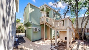 Une maison à deux étages avec un escalier en bois menant à un balcon, entourée d’une cour clôturée et d’arbres.