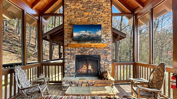 A cozy outdoor deck with a stone fireplace, wicker furniture, and a view of the surrounding forest.
