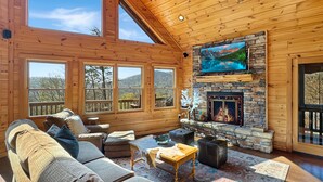 A cozy cabin interior with a stone fireplace, wooden walls, and large windows offering scenic views.