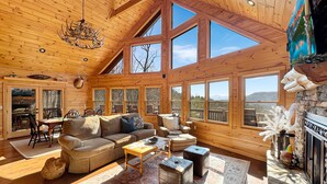 A spacious cabin with a high ceiling, large windows offering mountain views, a fireplace, and a cozy seating area.