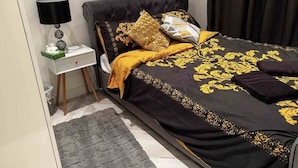 A bedroom with a black bedspread with gold patterns, a black and gold pillow, a white bedside table with a lamp, and a gray rug. - Immaculate 1-bed Apartment in the Heart of Chelsea (London)