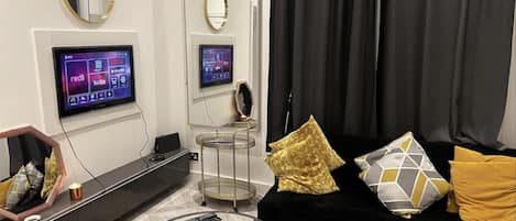 A modern living room with a black sofa, a glass coffee table, and a TV mounted on the wall.