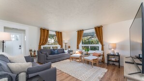 A modern living room with a grey sofa, a wooden coffee table, and a TV on a wooden stand. There are two side tables with lamps, a large window with curtains, and a view of greenery outside. - 5 Bedroom Home Downtown, Close to shops! (Chelan)
