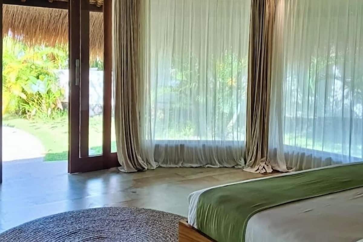 Une pièce avec un lit, de grandes fenêtres ornées de rideaux légers, une entrée surmontée d’un toit de chaume et une vue sur la verdure environnante.