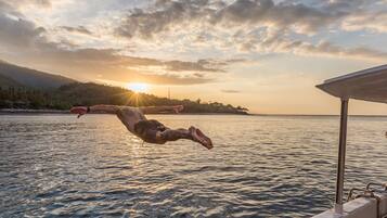Une personne plonge dans des eaux calmes au coucher du soleil, avec un bateau et une côte lointaine visibles.