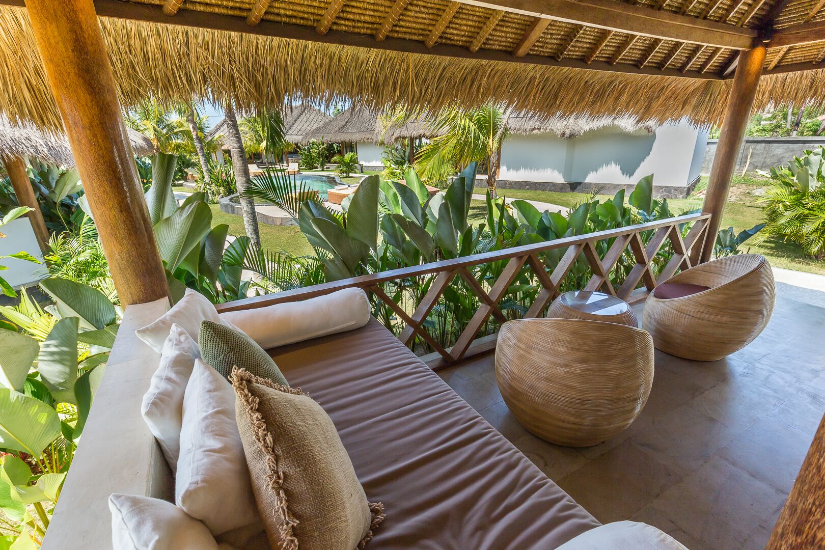 A balcony with a thatched roof, cushioned seating, and a view of a pool and lush greenery.