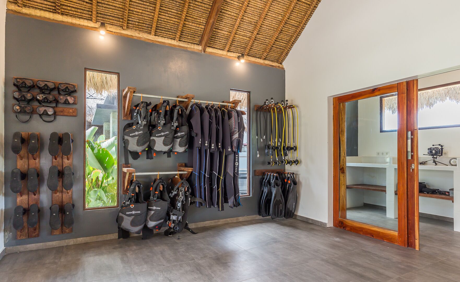 A room with a thatched ceiling, a wall-mounted rack of diving gear, and a wooden door leading to another room.