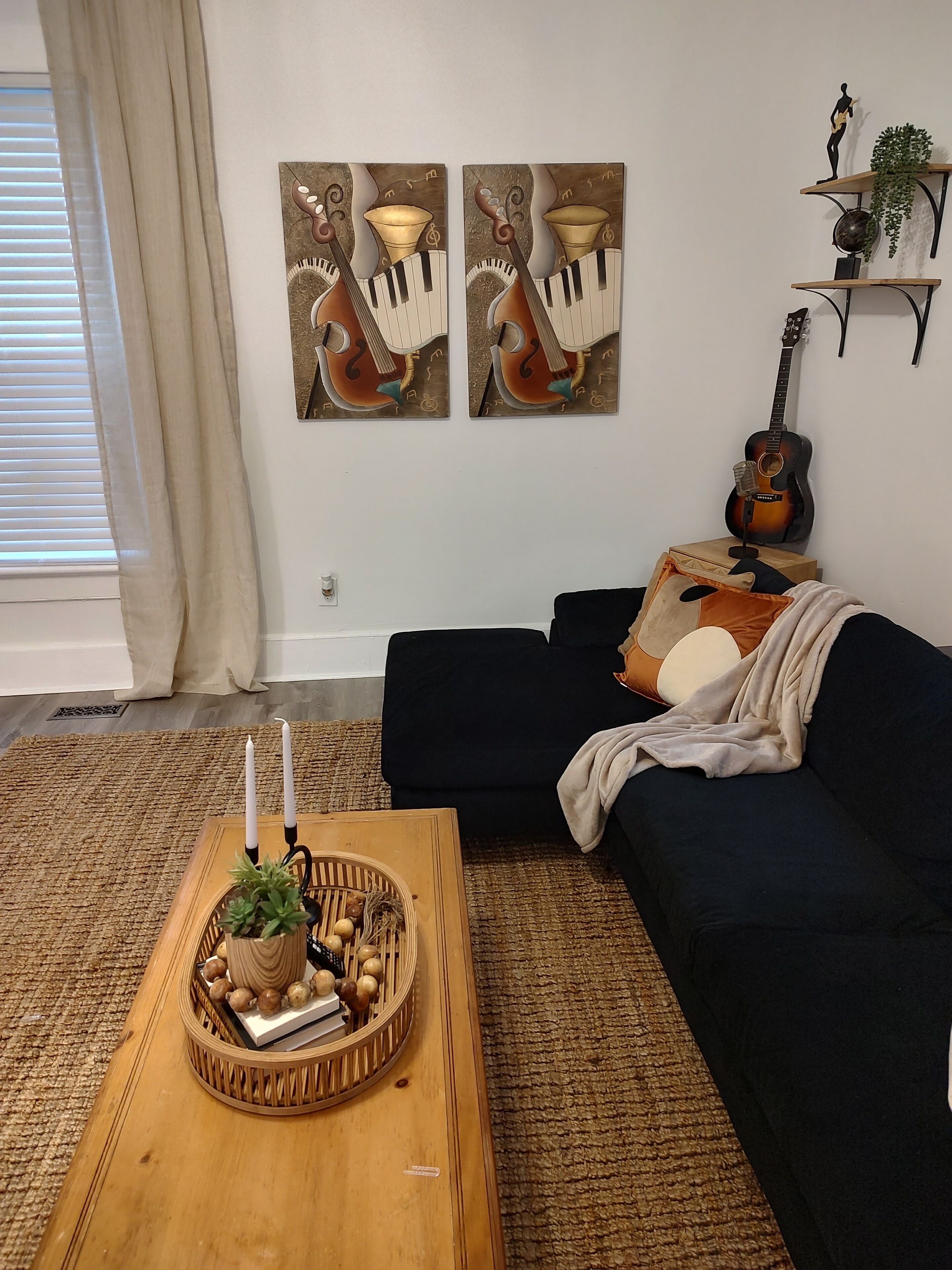 A living room with a dark blue sofa, a wooden coffee table with candles and a plant, and two wall-mounted paintings of musical instruments.