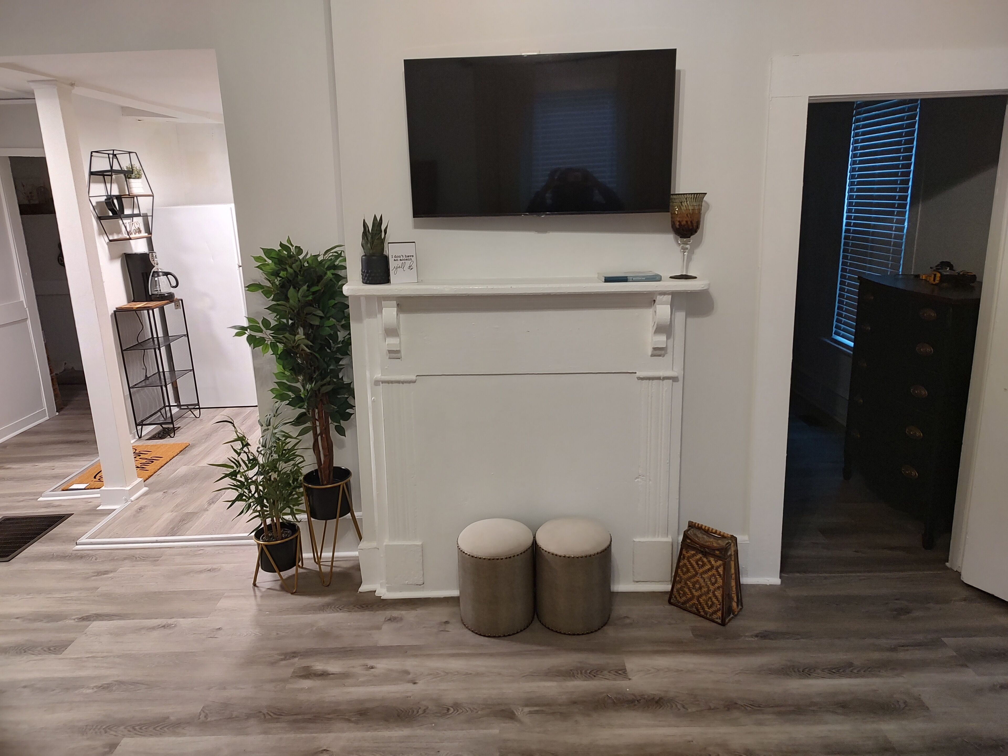 Un salon avec une cheminée blanche, un téléviseur à écran plat fixé au-dessus, deux poufs ronds et une plante en pot.