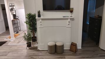Un salon avec une cheminée blanche, un téléviseur à écran plat fixé au-dessus, deux poufs ronds et une plante en pot.