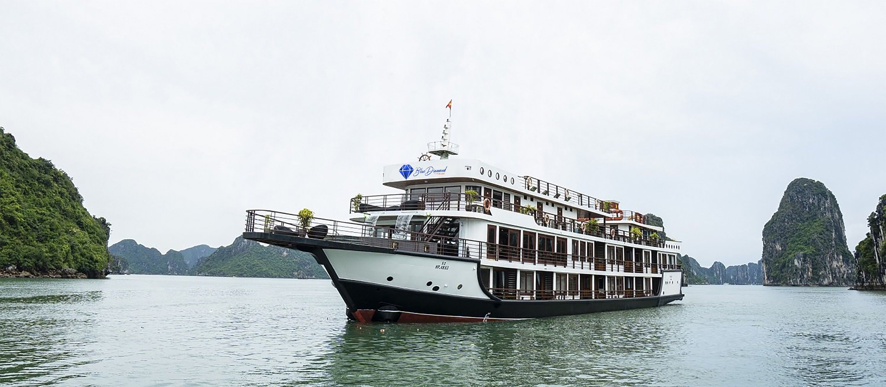 Ein großes, mehrstöckiges Boot mit weiß-schwarzer Farbgebung, genannt 'Blue Diamond', schwimmt auf dem Wasser vor dem Hintergrund grüner, gebirgiger Inseln.