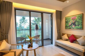 Een kamer met een bank, stoelen, een klein tafeltje met een vaas, en een balkon met uitzicht op palmbomen.