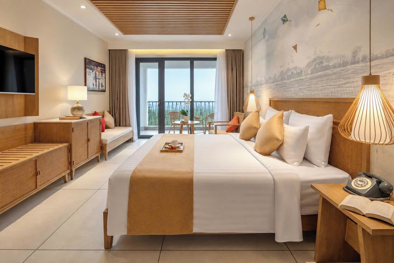 A hotel room with a bed, bedside table, lamp, and a view of the outdoors.