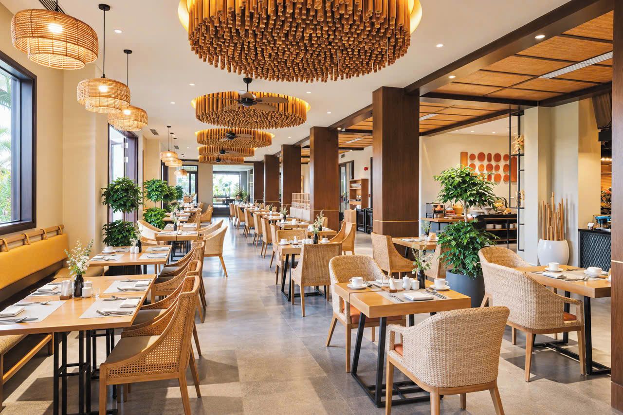A spacious dining area with multiple tables and chairs, large hanging light fixtures, and potted plants.