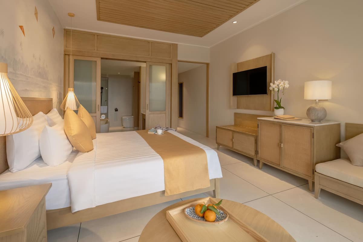 Une chambre d’hôtel moderne avec un grand lit, un téléviseur fixé au mur, une armoire en bois et une petite table sur laquelle se trouve un plateau de fruits.