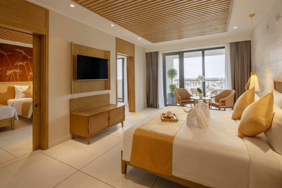 Une chambre d’hôtel moderne dotée d’un grand lit, d’un téléviseur fixé au mur, d’une tête de lit en bois, d’un coin salon avec des fauteuils et d’un balcon offrant une vue.