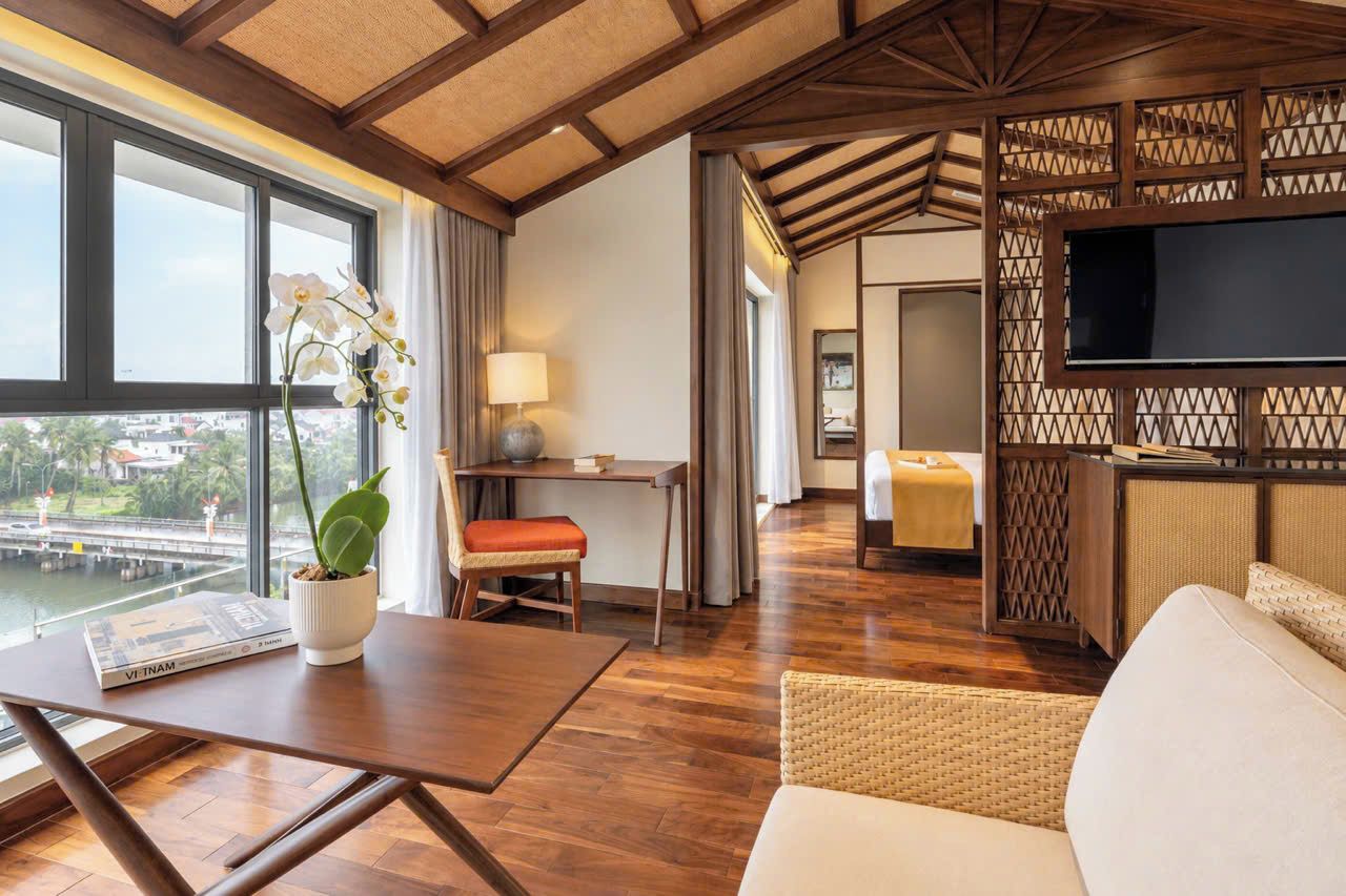 Habitación de hotel con piso de madera, zona de comedor con mesa y sillas, sofá, televisión y vista a un puente y al agua.