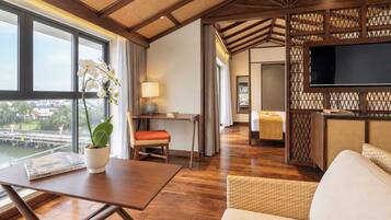 Habitación de hotel con piso de madera, zona de comedor con mesa y sillas, sofá, televisión y vista a un puente y al agua.