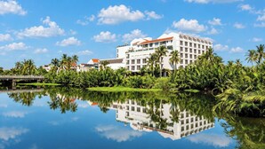 Un hôtel qui se reflète dans les eaux calmes d’un plan d’eau, entouré de verdure et de palmiers.