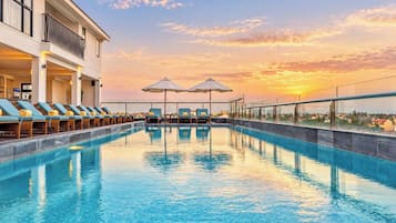 Una piscina con sillones de descanso y sombrillas, un edificio con balcones y vistas al atardecer.