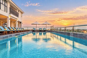 Une piscine avec des chaises longues et des parasols, un bâtiment doté de balcons et une vue sur le coucher de soleil.