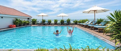 Piscina con sillones de descanso y sombrillas, rodeada de vegetación.