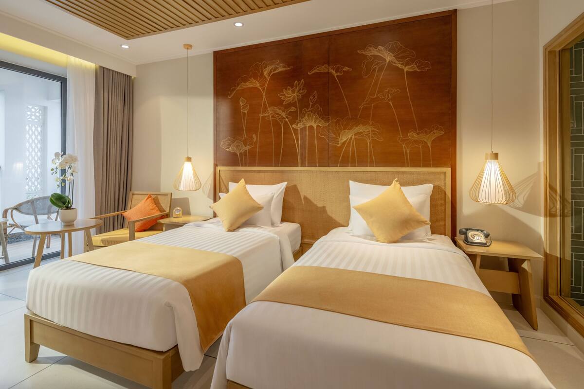 Une chambre d’hôtel avec deux lits, une tête de lit en bois, un mur décoré de motifs floraux, une petite table avec une lampe et une plante en pot.