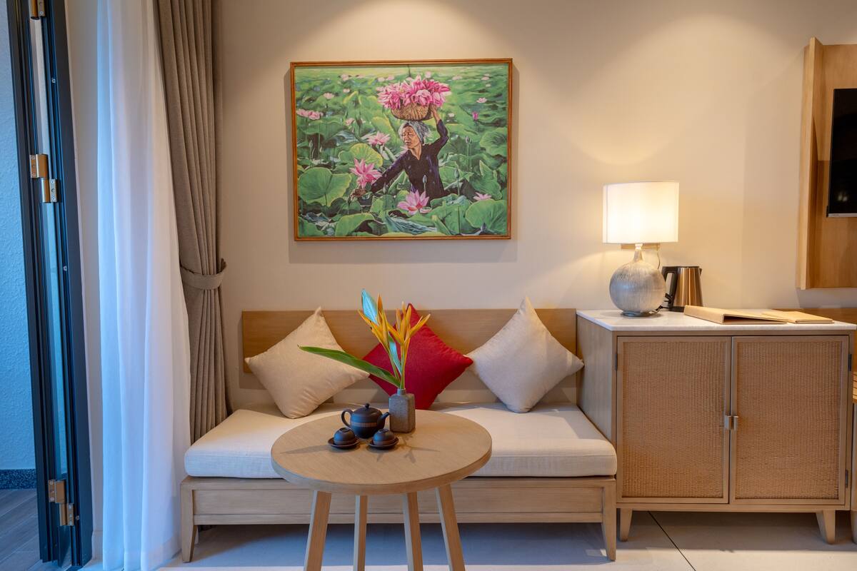 Une chambre d’hôtel moderne, dotée d’un canapé, d’une petite table ronde avec une théière et des fleurs, d’une armoire et d’un tableau représentant un étang de lotus.