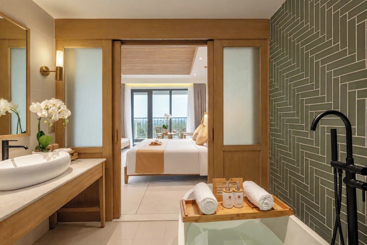 A hotel room with a large bed, a wooden bench with towels, a sink, and a mirror.