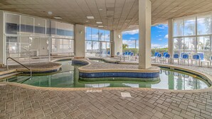 An indoor swimming pool with tiled edges, a brick floor, and a view of palm trees through large windows. - Carolinian 931 (Myrtle Beach)