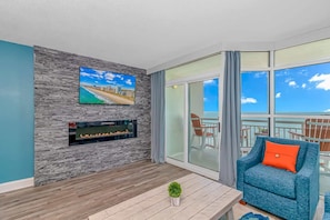 A balcony with a view of the sea, a TV mounted on a textured wall, a fireplace, and a blue sofa with an orange pillow.