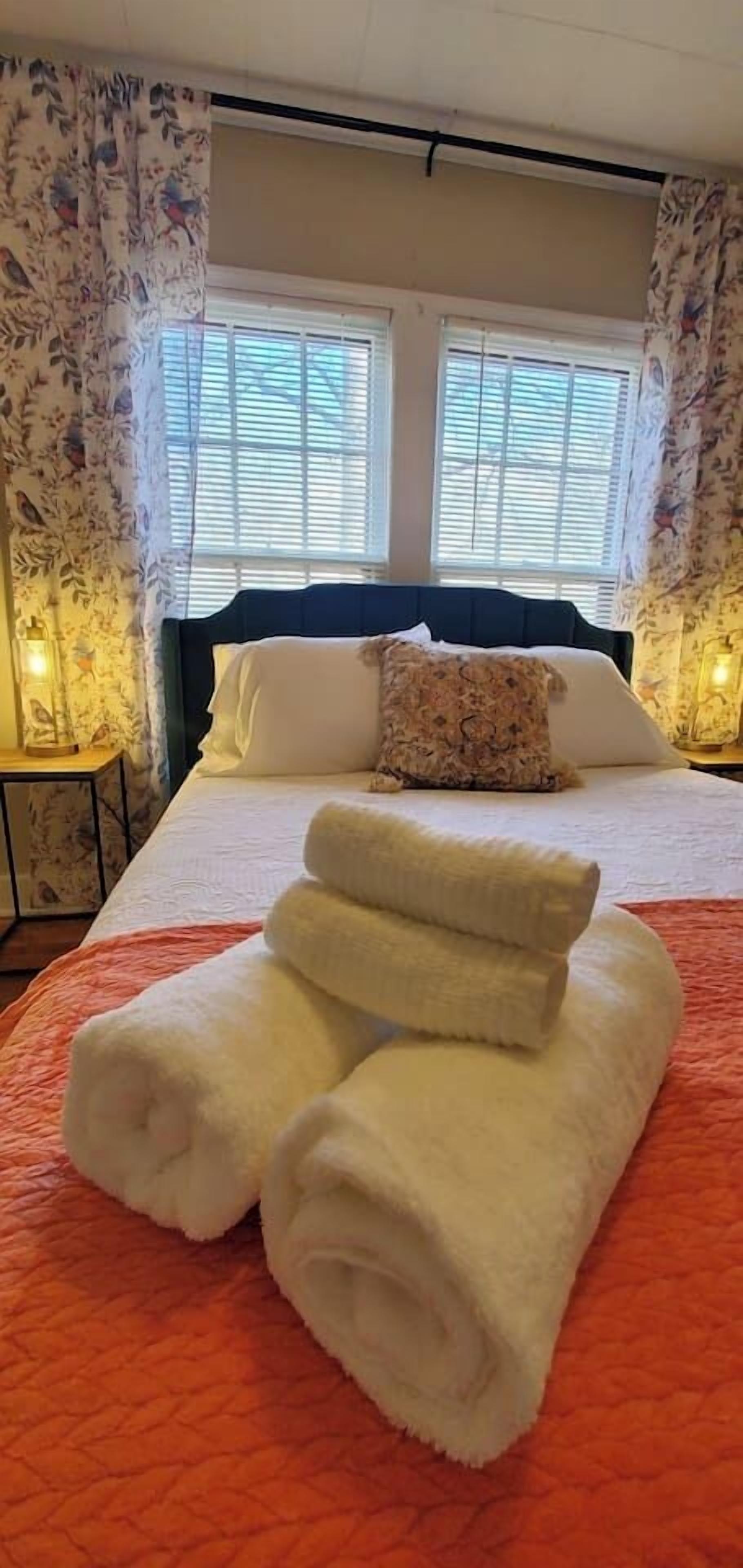A neatly made bed with white linens and a folded towel in the center, a floral-patterned curtain, and a window with blinds.