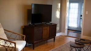 A living room with a television on a wooden cabinet, a wicker chair, a small round table, and a door leading to another room.