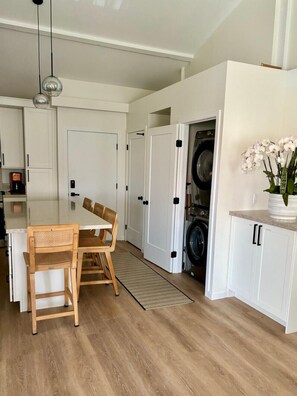 Una cocina moderna con armarios blancos, un comedor con mesa y sillas de madera, y una lavadora al fondo.