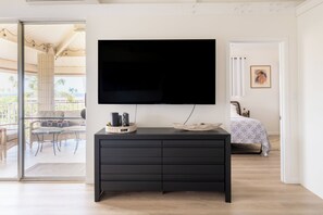 Un salon moderne avec un téléviseur à écran plat fixé à un meuble noir, une table en verre avec des chaises, et une vue sur un balcon aménagé avec du mobilier d’extérieur.